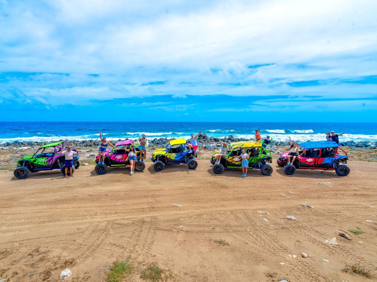 Colorful ATVs parked on beach, people standing, ocean in background.