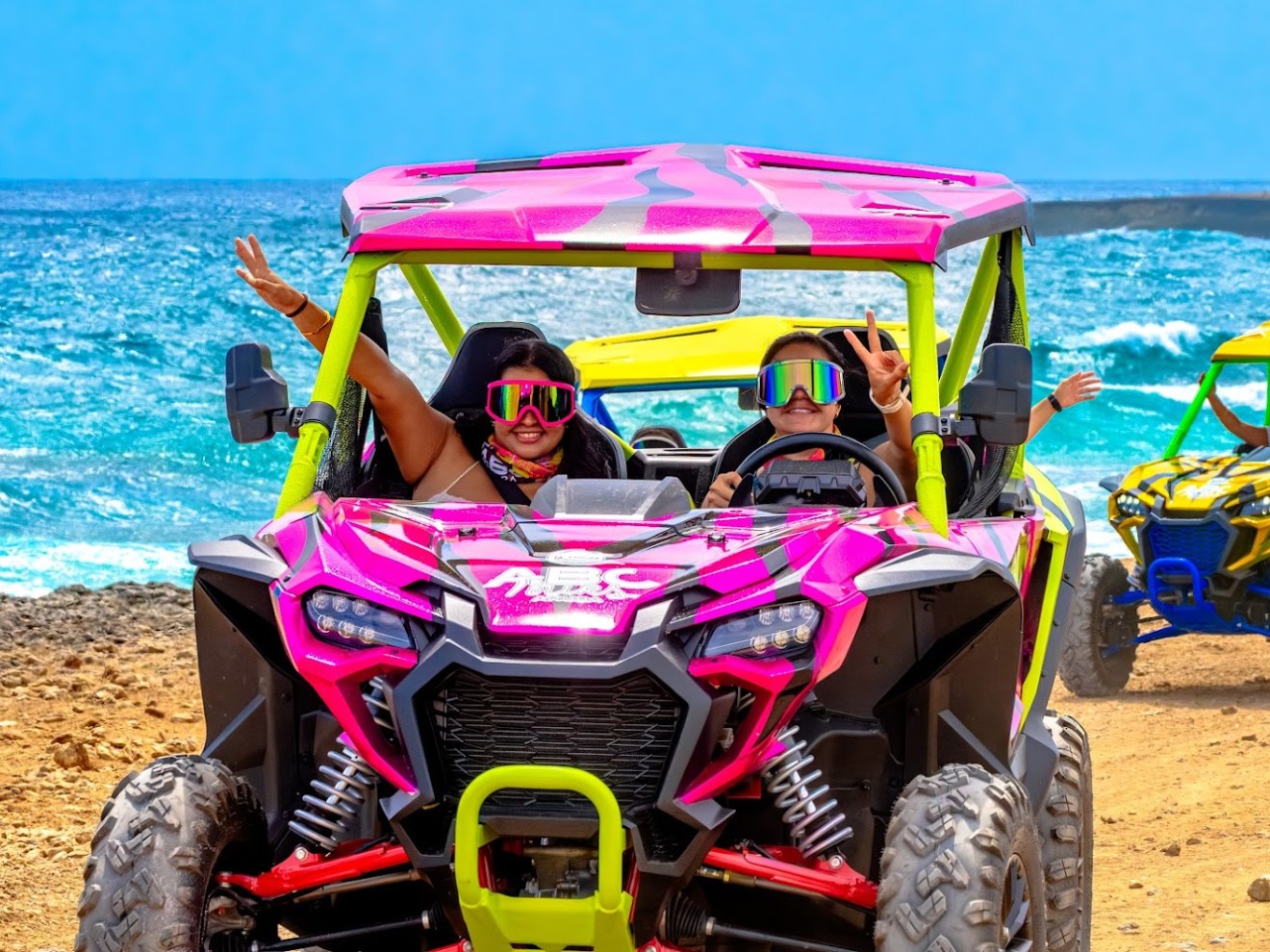 Two people in a pink off-road vehicle near the ocean, both wearing goggles.