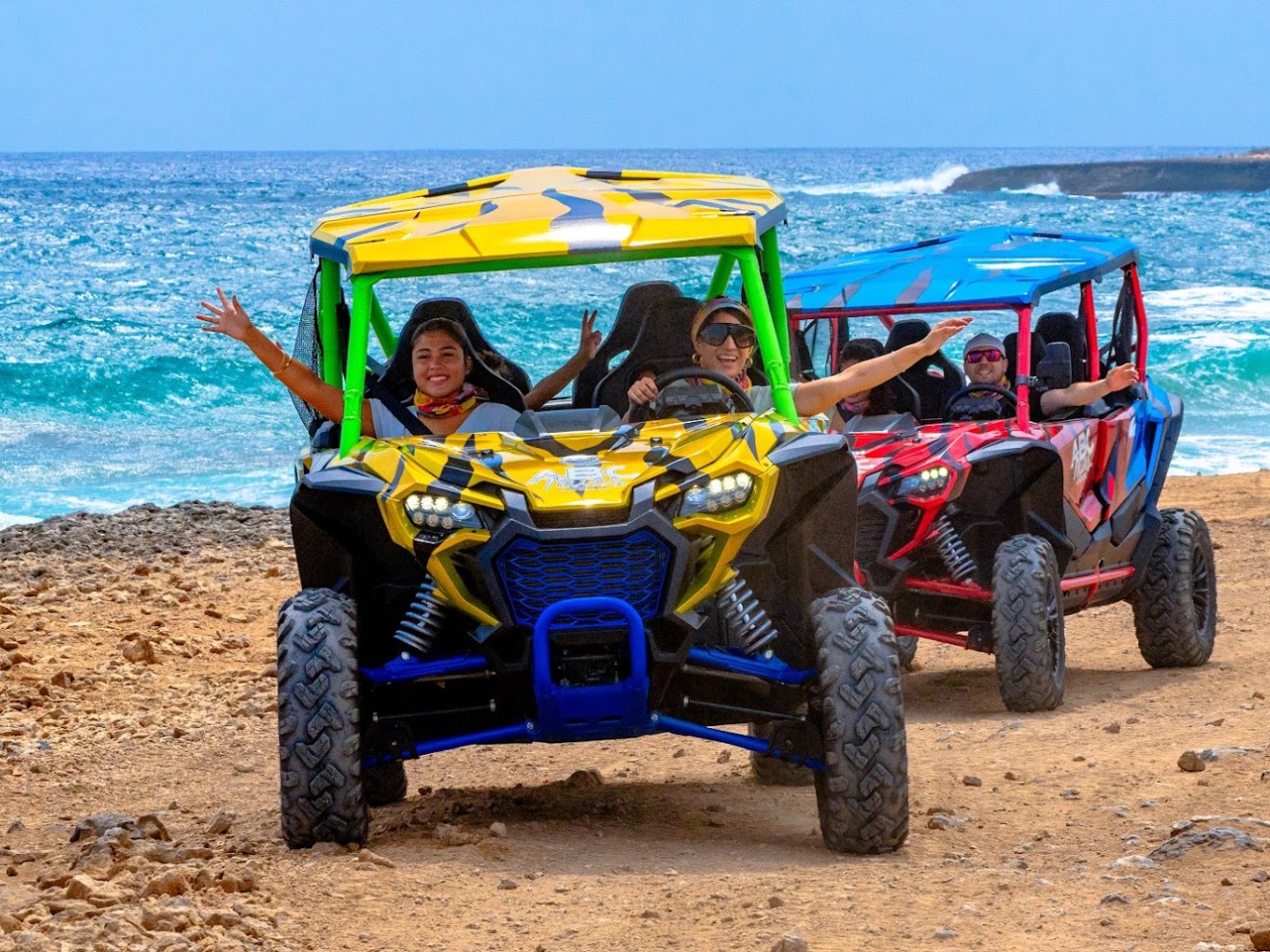 Two dune buggies with happy passengers drive along a rocky beach.