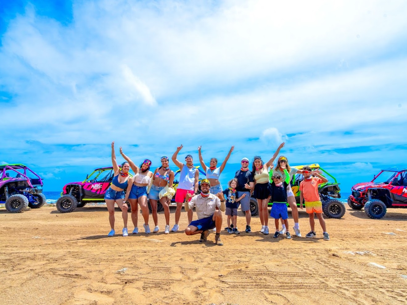 Group of people posing with colorful off-road vehicles on sandy terrain under a blue sky.