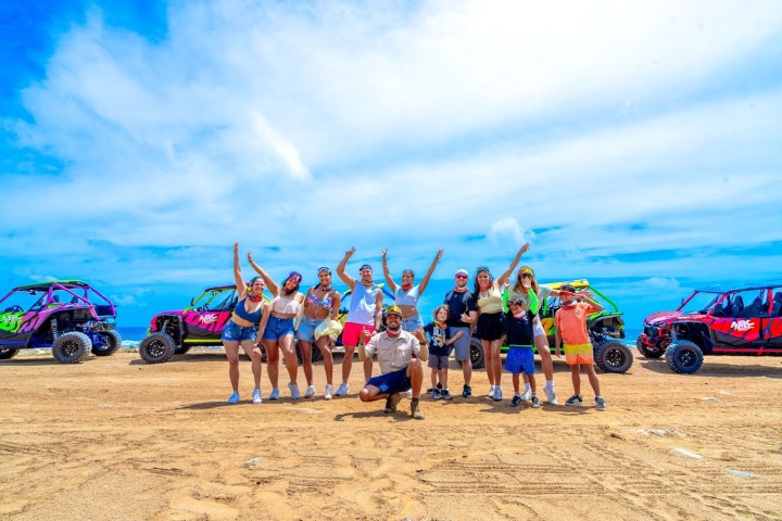 Group of people posing with colorful off-road vehicles on sandy terrain under a blue sky.