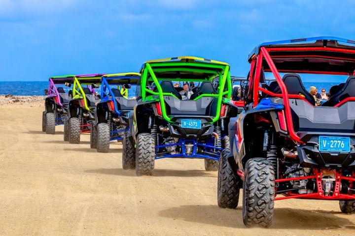 Colorful off-road vehicles driving in a line on a sandy path under a blue sky.