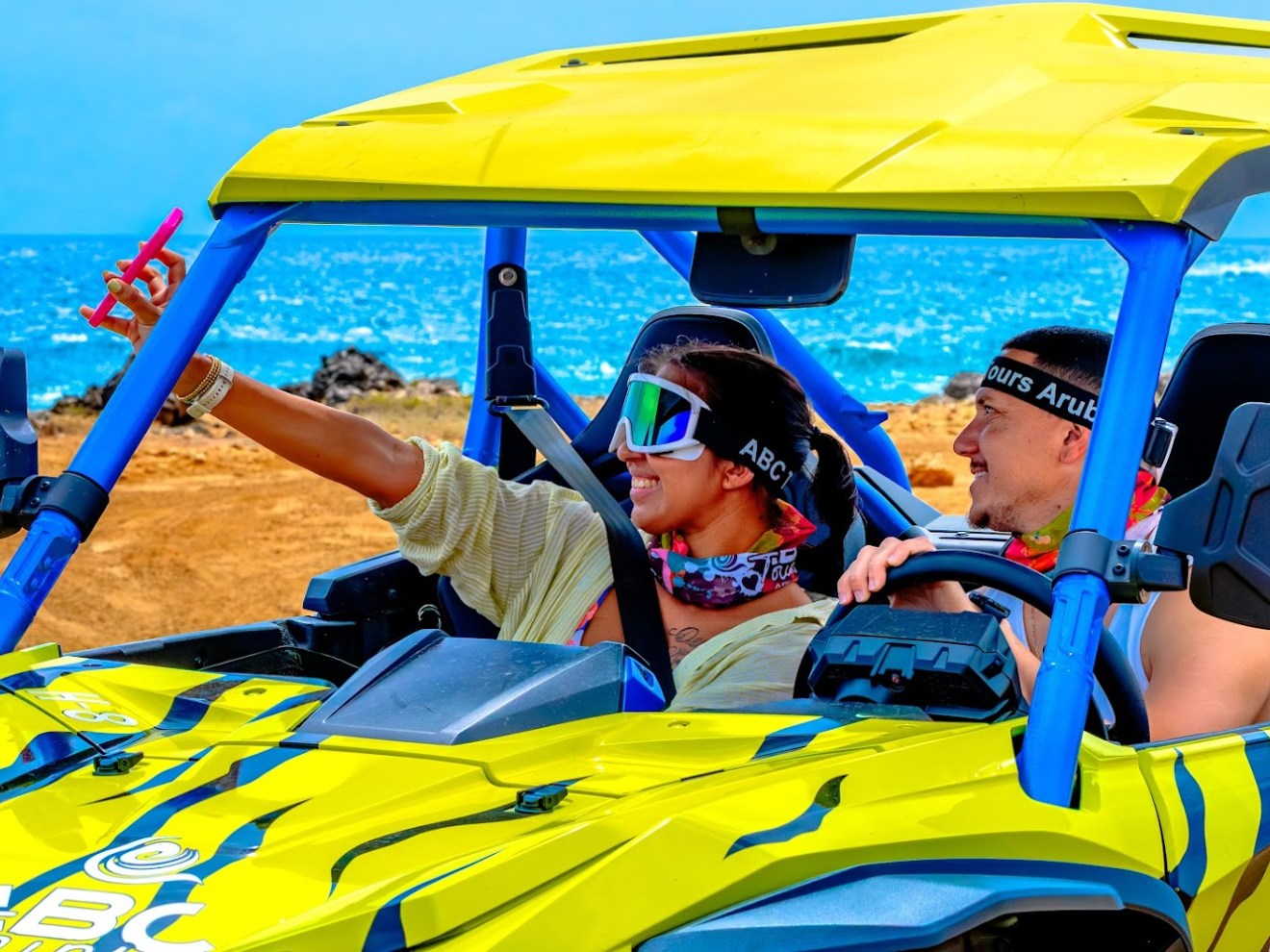 Two people in a yellow off-road vehicle by the sea, one taking a selfie.