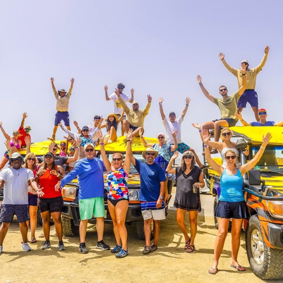 Group posing with arms raised near SUVs by a lighthouse.